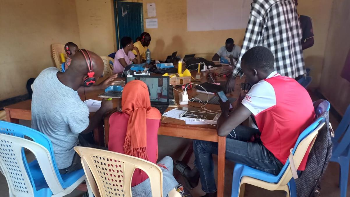 A group of people sit around a wooden table in a classroom, working on laptops and phones, with notebooks, cables, and containers scattered across the table.