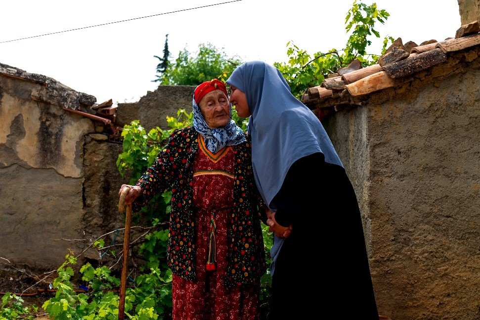 Beni Maoush Kabyle, Kabyle elder stands beside Fella Lahmar, DEPA researcher who leans in to speak with her, both outdoors near a stone wall and grapevines.