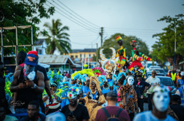 Carnival participants in colourful costumes and masks moving along a road lined with buildings and palm trees, including stilt walkers in the background.