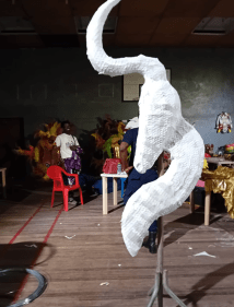 A large white, horn‑shaped sculptural piece sits on a metal stand in the foreground. Behind it, several people work around tables covered with colourful fabrics and art materials in a bright indoor workshop space with wooden floors