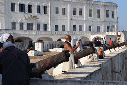Visitors stand near a row of rusted cannons outside a large whitewashed coastal fort, with people taking photos and exploring the historic site.