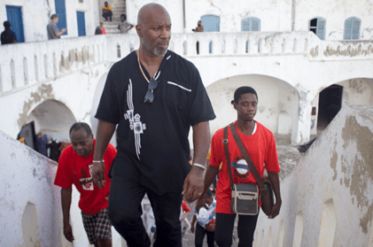 People walk up a staircase at cape Coast Castle, a whitewashed historic building with arched architecture and blue doors