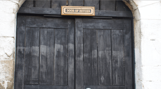 A pair of large, dark wooden doors set in a stone archway with a sign above them that reads “Door of Return.”