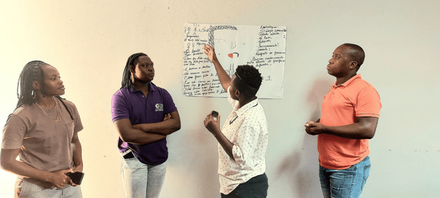 Four people at the workshop, stand in front of a large sheet of paper taped to the wall. One person points to the paper while presenting, and the others watch attentively. The sheet shows a simple drawing of a face and a handwritten list of communication and respect guidelines.