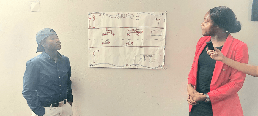 Two people stand beside a large poster labelled “GRUPO 3,” which shows a hand‑drawn street scene with cars, motorbikes, traffic lights, and a bus stop, showing places they have been accosted as trans people