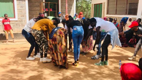 A group of people stand in a circle outdoors, leaning in with their hands extended toward the centre during a group activity, with others watching nearby in a courtyard surrounded by stone and white buildings.