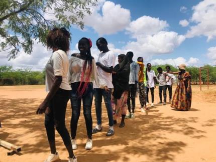A line of people stand outdoors on sandy ground, participating in an activity together, with sparse trees in the background and a teacher wearing a brightlytraditional outfit at the end of the line.