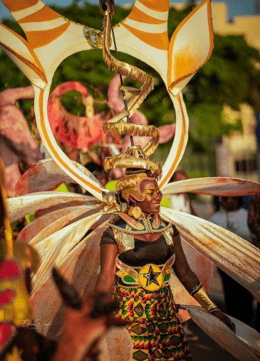 A female performer wearing a large, colourful carnival costume with wing‑like extensions, a dramatic spiral headpiece, and a patterned skirt, surrounded by other participants in a festive parade.