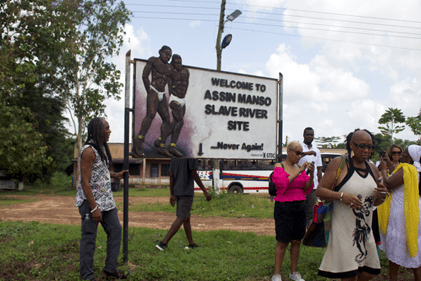 A group of people stand near a large sign reading “Welcome to Assin Manso Slave River Site – Never Again,” with an illustration of two shackled figures, set outdoors with greenery, a building, and a parked bus in the background.