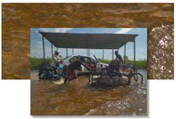 A metal-roofed shelter stands in a flooded area, with a white car, motorcycles, and a tricycle partially submerged beneath it, while several people navigate the high water around the vehicles.