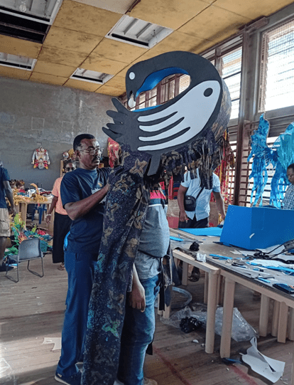 Johnson in the workshop adjusting a large, stylised bird‑shaped headpiece, surrounded by colourful costume materials and tools.