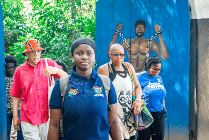 People walk through an outdoor entrance featuring a large mural of a man breaking free from chains, surrounded by lush greenery.
