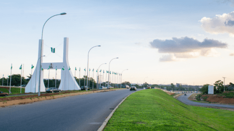 A large white gateway stands over a road lined with Nigerian flags, with cars approaching under a partly cloudy sky.